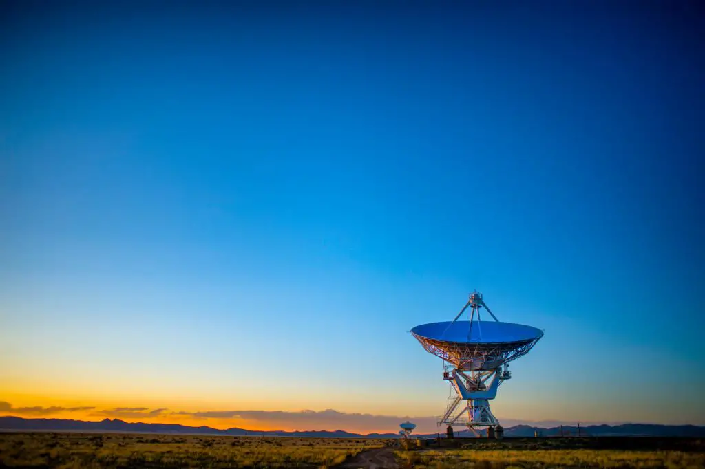 A large radio telescope dish stands solitary against the backdrop of a twilight sky, with hues of orange and blue over the New Mexico landscape. This is the Very Large Array in New Mexico.