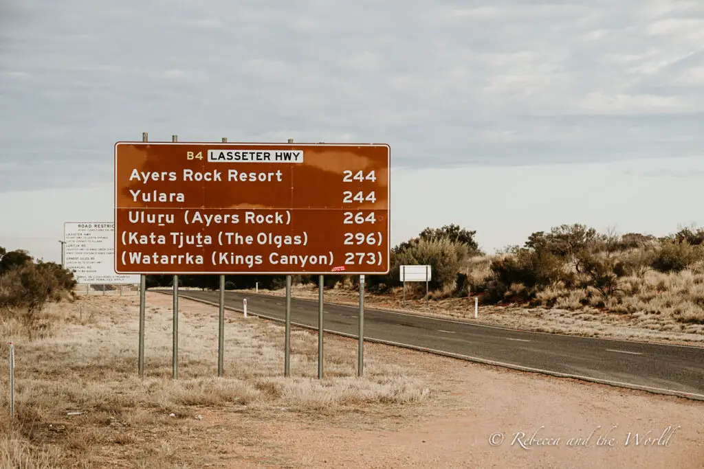 A roadside sign indicating directions and distances to various locations, including Ayers Rock Resort, Yulara, Uluru (Ayers Rock), Kata Tjuta (The Olgas), and Watarrka (Kings Canyon), against a backdrop of a flat, arid landscape. Driving distances are long on a Red Centre Way road trip.