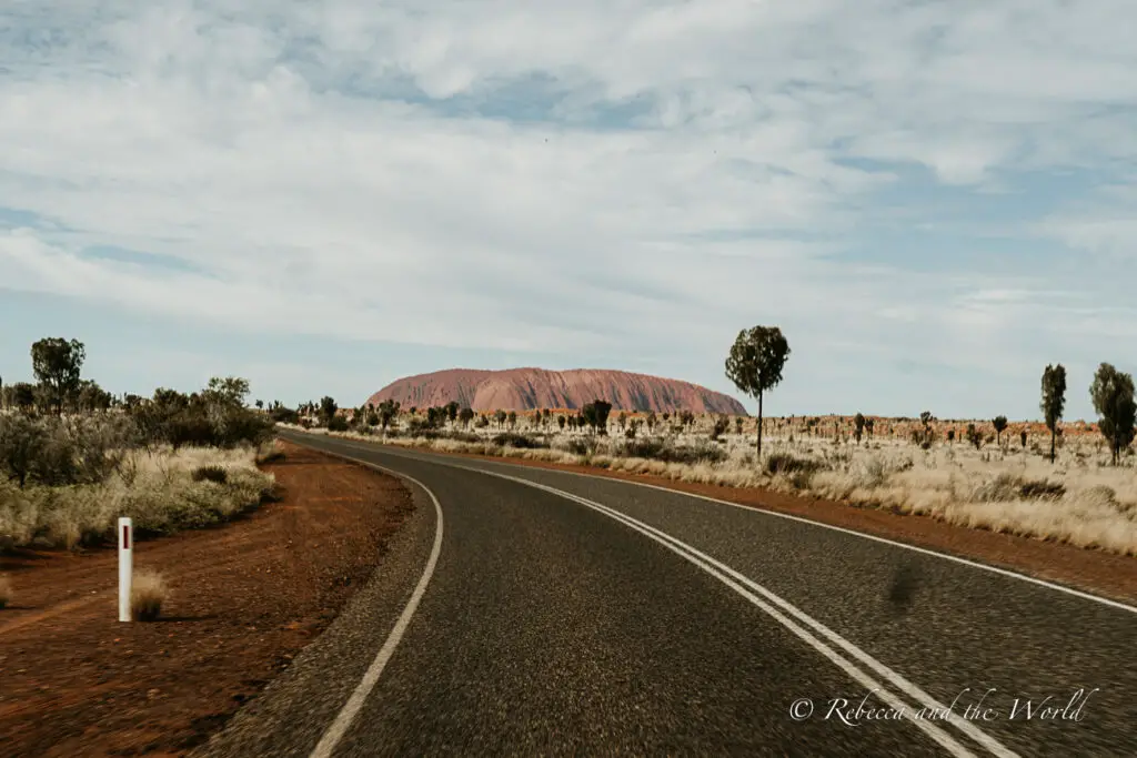 A road in the Australian Outback leading towards Uluru, visible in the distance with its distinctive red colouring, under a sky with light cloud cover. It's hard to forget the first time seeing Uluru rising up before you.