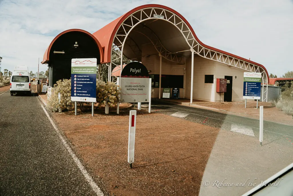 The entrance of Uluru-Kata Tjuta National Park with a sign that includes park rules and regulations. There are camper vans visible, indicating the popularity of road trips to this destination.