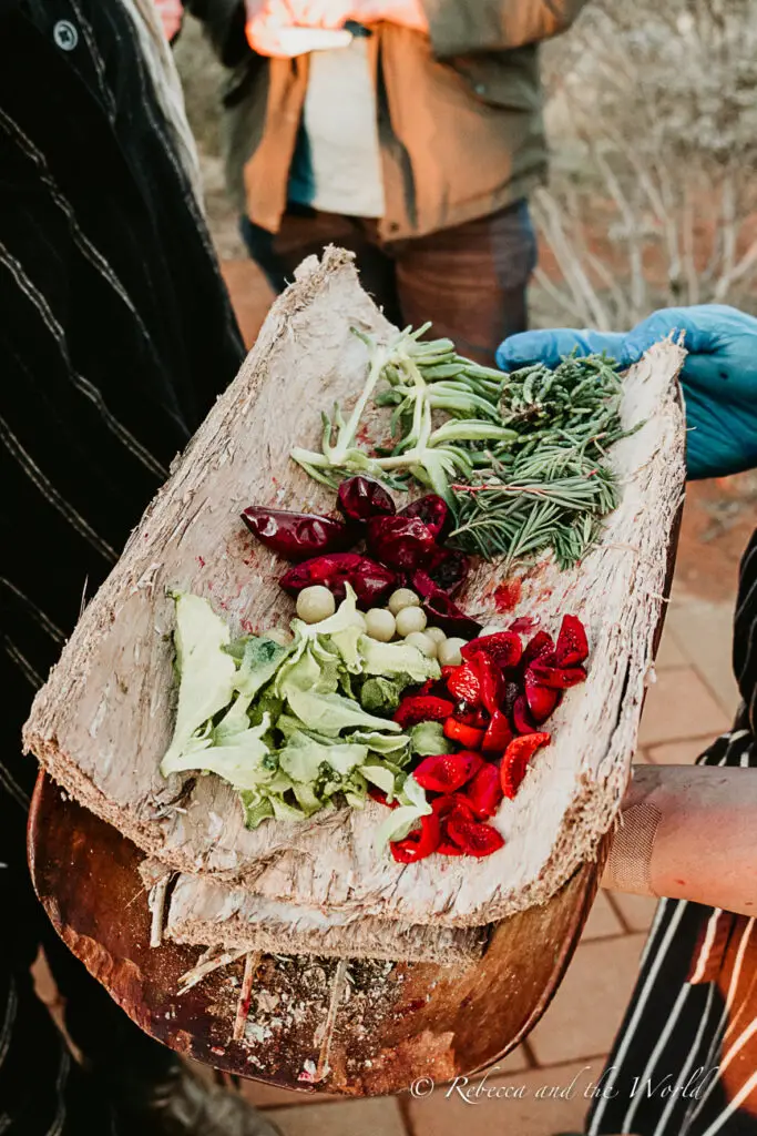 A wooden plank serving as a rustic cutting board with a selection of fresh vegetables and herbs, held by someone wearing a blue glove, with people in the background.