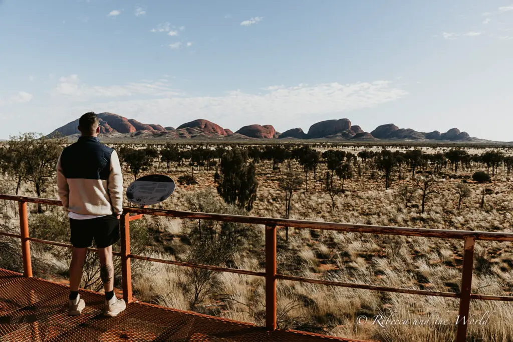 A man - the author's husband - standing on an elevated viewing platform overlooking the Kata Tjuta (The Olgas) formations in the distance, under a clear blue sky. Kata Tjuta means “many heads” in Pitjantjatjara language. Kata Tjuta is a must-see on an Alice Springs to Uluru road trip.