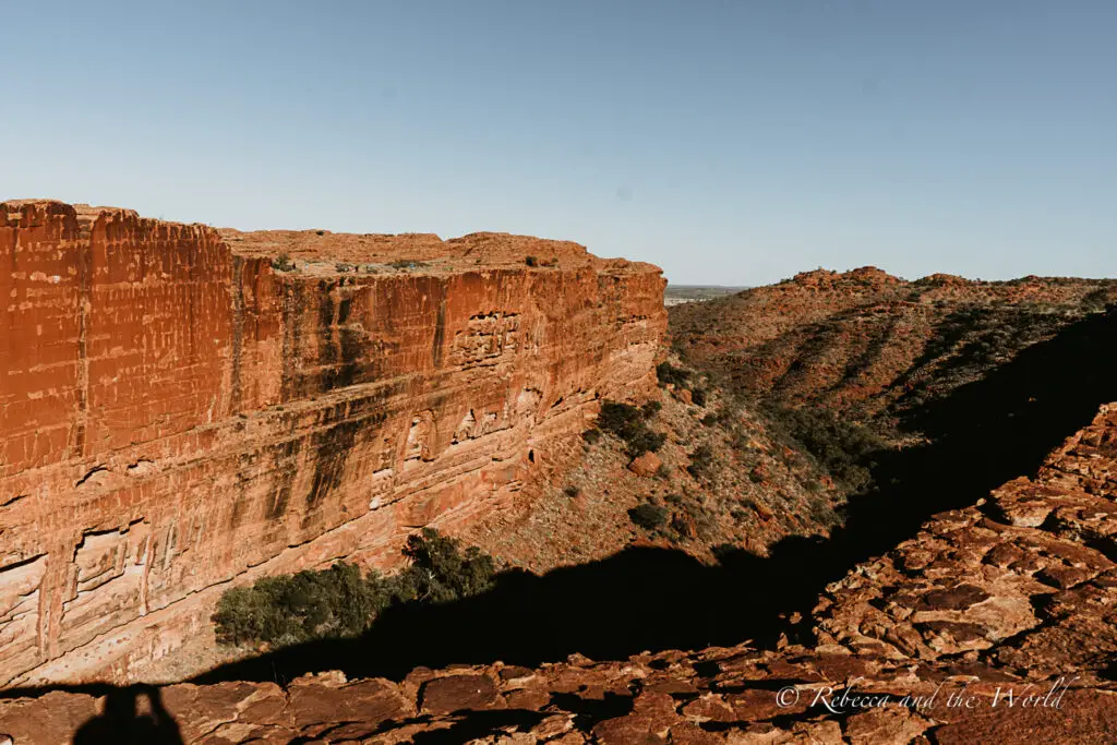 A panoramic view of a grand, layered red rock cliff under a clear blue sky. The cliff's geological striations are visible, and it overlooks a rugged canyon with sparse vegetation. This is part of the Kings Canyon Rim Walk in Watarrka National Park, Northern Territory.