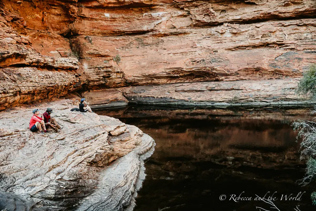 A group of people sitting on a rock ledge by a waterhole in a desert setting, enjoying a peaceful break during their journey. The Garden of Eden on the the Kings Canyon Rim Walk - one of my recommended must-dos on a Red Centre Way road trip.