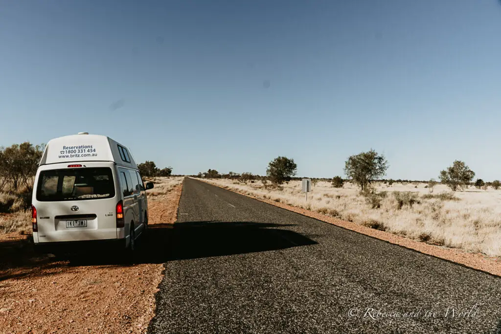 A white rental van parked on the side of a desolate asphalt road in the Northern Territory, with dry grassland and scattered shrubs on either side. The sky is clear and blue.