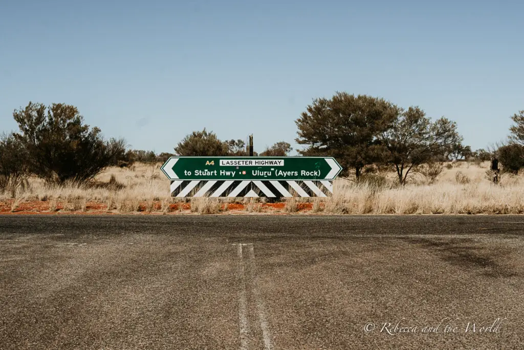 A roadside image with a direction sign showing "A4 Lasseter Highway" pointing to the right towards "Uluru (Ayers Rock)" and to the left towards "Stuart Hwy." The sign is in the foreground with sparse Outback vegetation in the background under a clear blue sky.