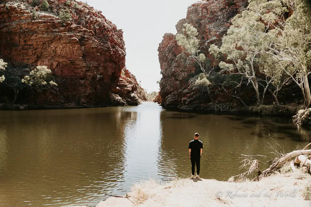 A calm gorge framed by high red rock cliffs and green foliage. A solitary figure - the author's husband - stands at the water's edge, looking into the distance, creating a serene and reflective scene. This is Ellery Creek Big Hole, one of the best places to visit in the Northern Territory.