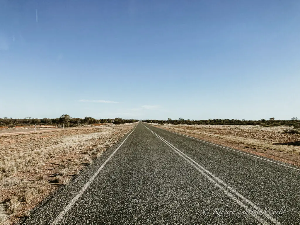 A straight, empty road cutting through the Australian Outback with wide-open skies and flat, arid landscape stretching into the distance. The roads are long and flat on an Alice Springs to Uluru road trip.