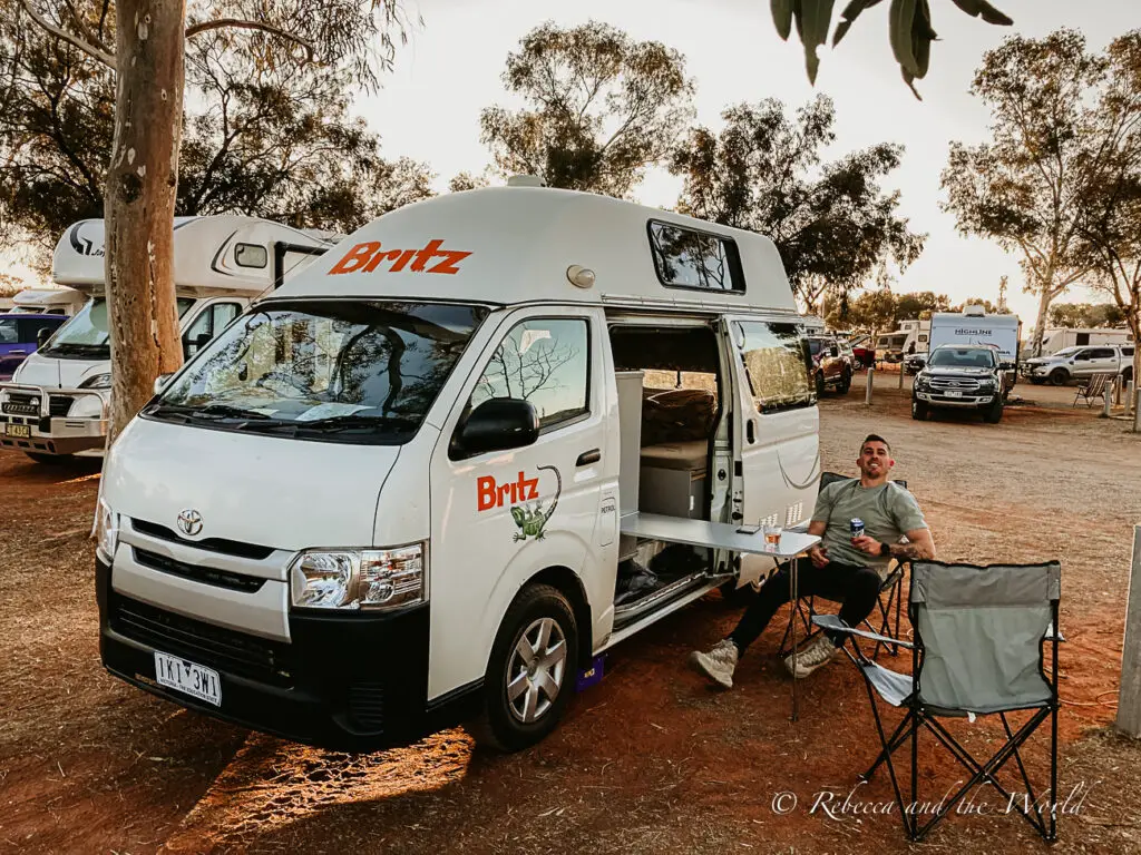 A camper van with the "Britz" logo parked at a campsite with trees around. A man - the author's husband - is seated outside on a camping chair next to a small table, smiling at the camera, enjoying the outdoor camping experience. Having a campervan for this Uluru self drive itinerary and Central Australia road trip will give you a lot more flexibility.