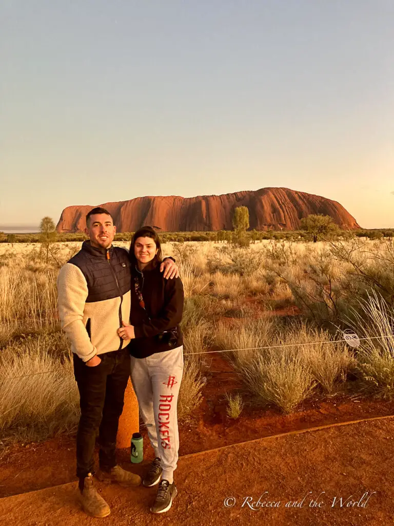 A couple - the author and her husband - posing for a photo with Uluru in the background. They are dressed in casual outdoor attire and are standing on a red dirt path among the grassy plains.