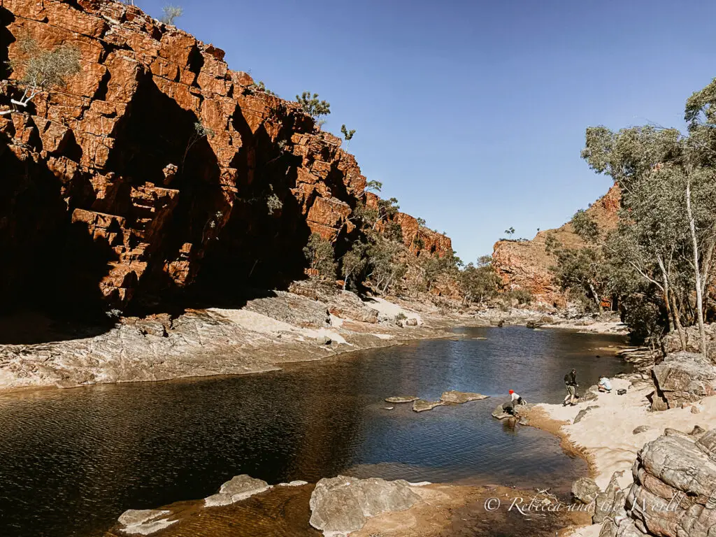 A serene waterhole nestled among red rock cliffs, with people enjoying the natural beauty and calm waters. Ormiston Gorge in the West MacDonnell Ranges (Tjorita).