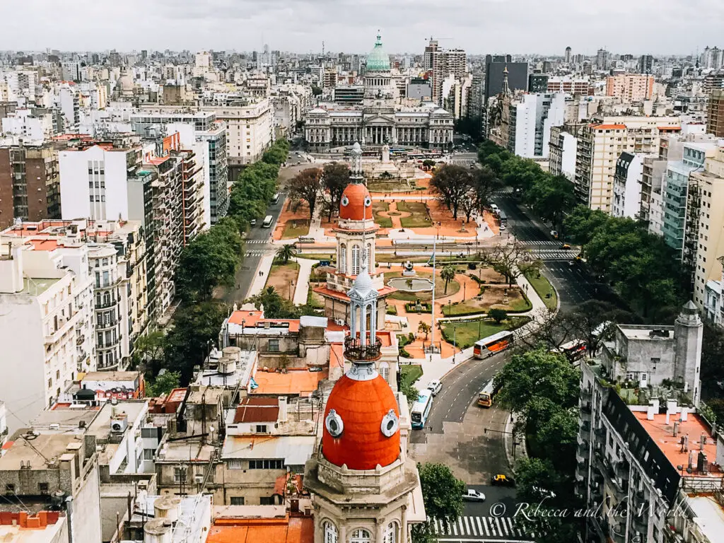An aerial view of Buenos Aires from Palacio Barolo, showing dense, varied architecture, a large green park in the centre, and a striking red-domed building in the foreground. The background shows a cloudy sky over the urban expanse.