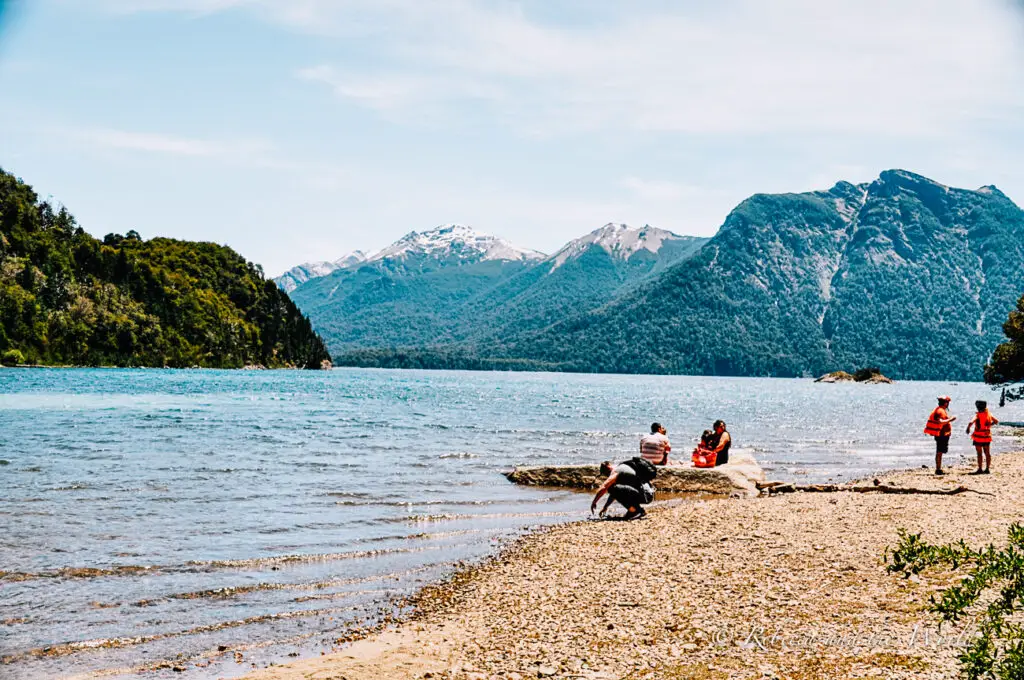 A shoreline near Bariloche with clear blue water, with people sitting and exploring, some wearing orange life vests. The background features majestic mountains with snowy peaks, under a bright blue sky.