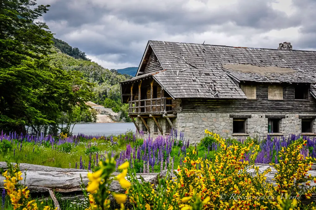 A rustic two-story stone house with a wooden balcony, surrounded by lush greenery and vibrant purple and yellow wildflowers. A tranquil lake and forested hills are visible in the background under a cloudy sky. This is some of the stunning scenery along the Ruta de los Siete Lagos - one of the best road trips in Argentina.