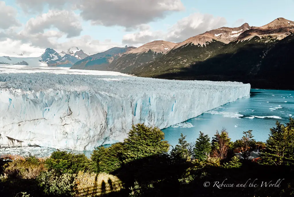 A panoramic view of Perito Moreno Glacier flowing into a lake. The glacier's surface is a vivid blue-white, and it is surrounded by dark green foliage and distant mountains under a partly cloudy sky. One of the best things to do in Argentina is go ice trekking on Perito Moreno Glacier.