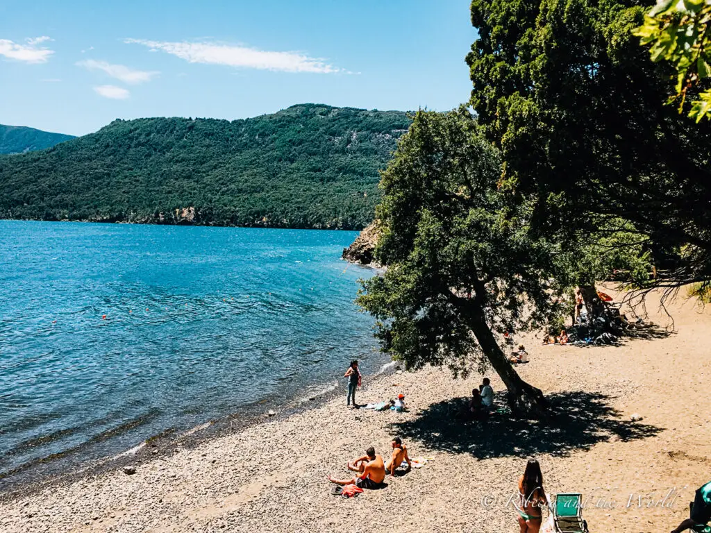 A handful of people sit on a rocky beach by a lake in Argentina. Some sit under the shade of a tree whiles others enjoy the full sunshine. The lake is near Bariloche in Argentina in summer.