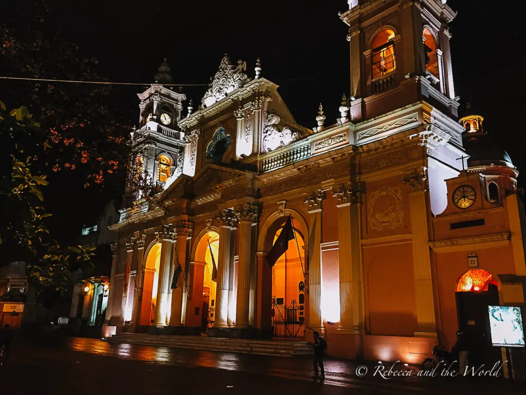 A grand, ornately decorated church in Salta illuminated at night, with its facade bathed in warm orange light against the dark sky. Its architectural details are highlighted, and a few people can be seen near the entrance. Salta is one of the many gorgeous cities in Argentina.