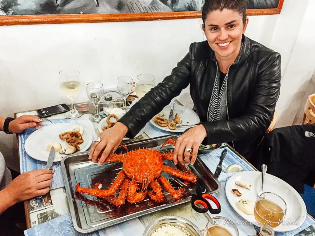 A woman (the author of the article) at a dining table preparing to eat a large, bright orange crab. Various dishes, glasses, and a smartphone are scattered on the table, capturing a lively mealtime atmosphere. Try Ushuaia's famous king crab!