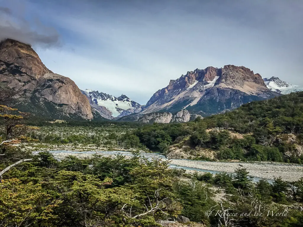 A rugged landscape in El Chalten, Argentina, featuring a mix of sharp and rounded mountain peaks, some with snowy crests. A creek winds through a valley with lush greenery under a partly cloudy sky. El Chalten is one of the best places to visit in Argentina for hiking.