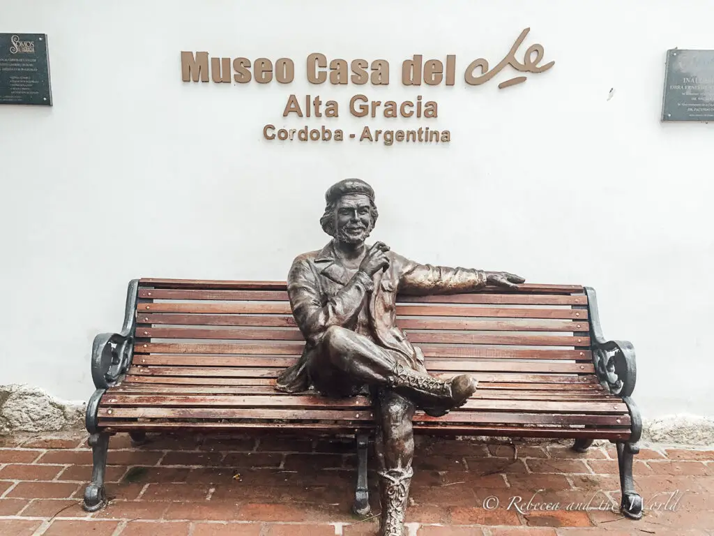 A bronze statue of a smiling Che Guevara sitting on a bench, which is part of the "Museo Casa del Che" in Alta Gracia, Córdoba, Argentina, as indicated by a sign above. In Cordoba in Argentina you can visit Che Guevara's childhood home, which is now a museum.