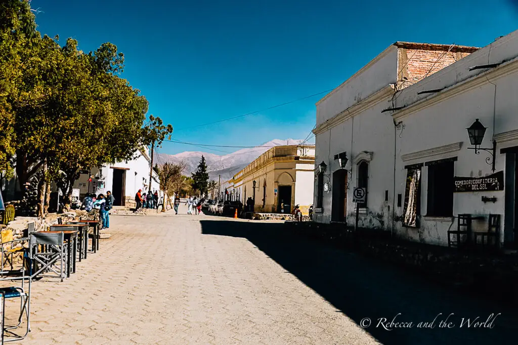 A street view of Cachi in Northwest Argentina shows a paved road, low buildings and chairs and tables on the side of the street. There are mountain ranges in the background.