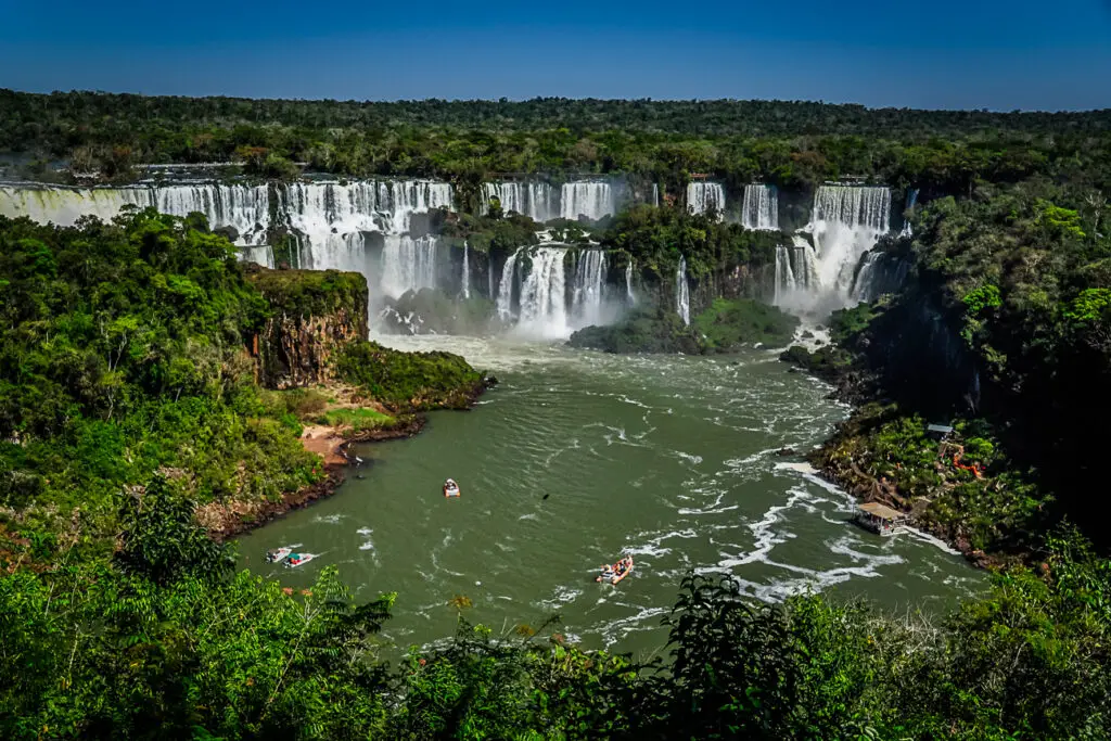A view from above shows Iguazu Falls - one of the best honeymoon in Argentina destinations - with several boats on the water below the the falls. The waterfalls are surrounded by lush green rainforest.