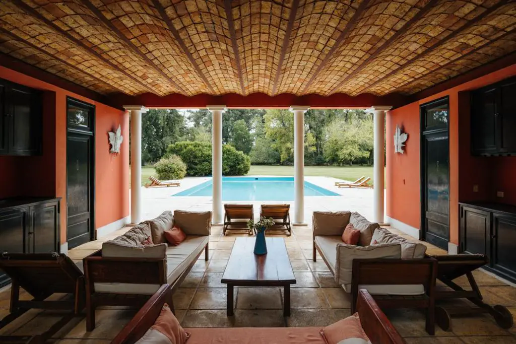 A covered outdoor patio with comfortable seating overlooking a swimming pool, with open views to a well-manicured lawn. La Bandada is a beautiful estancia near Buenos Aires, Argentina, known for its polo and outdoor activities.