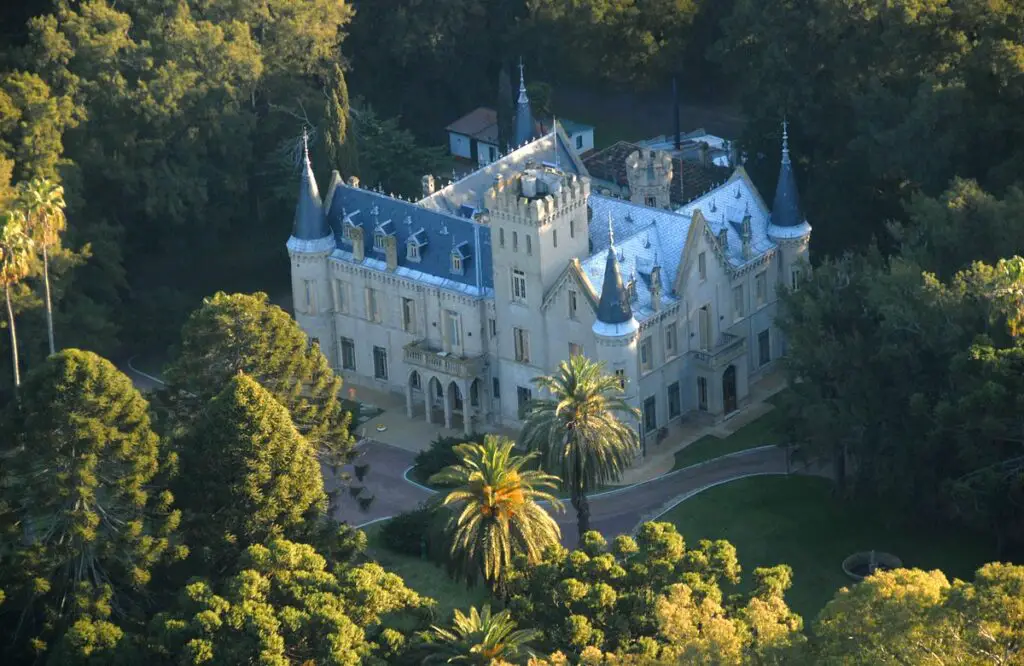 An aerial view of an ornate, castle-like building amidst lush greenery, with tall palm trees and a clear sky above. The property is Estancia La Candelaria, one of the most luxurious estancias near Buenos Aires.