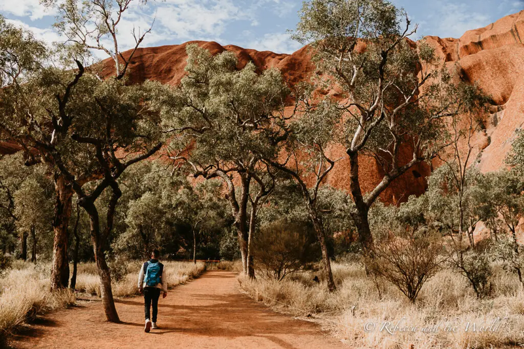 A path framed by green leafy trees leading to Uluru, with a person in a blue backpack walking away from the viewer. The Uluru Base Walk is one of the best things to do at Uluru to get up close to the monolith.