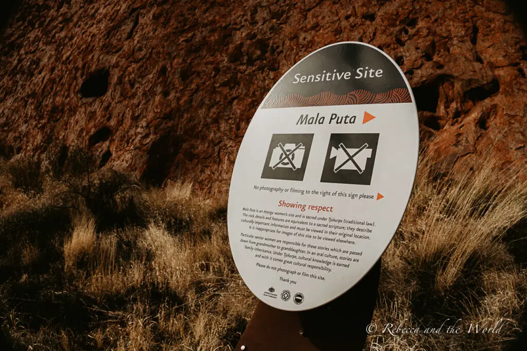 A close-up of a sign reading "Sensitive Site - Mala Puta" with symbols indicating no photography or filming to the right, set against the textured backdrop of Uluru's rock surface. When visiting Uluru, be respectful of the areas where you can't take photos so that you can protect sacred areas.