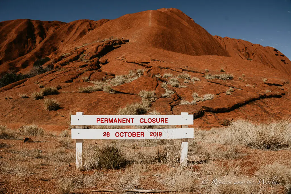 A white sign with red lettering stating "PERMANENT CLOSURE 26 OCTOBER 2019" in front of Uluru, indicating the closing of climbing activities on the rock. You can no longer climb Uluru, a move made out of respect for the local Anangu people.