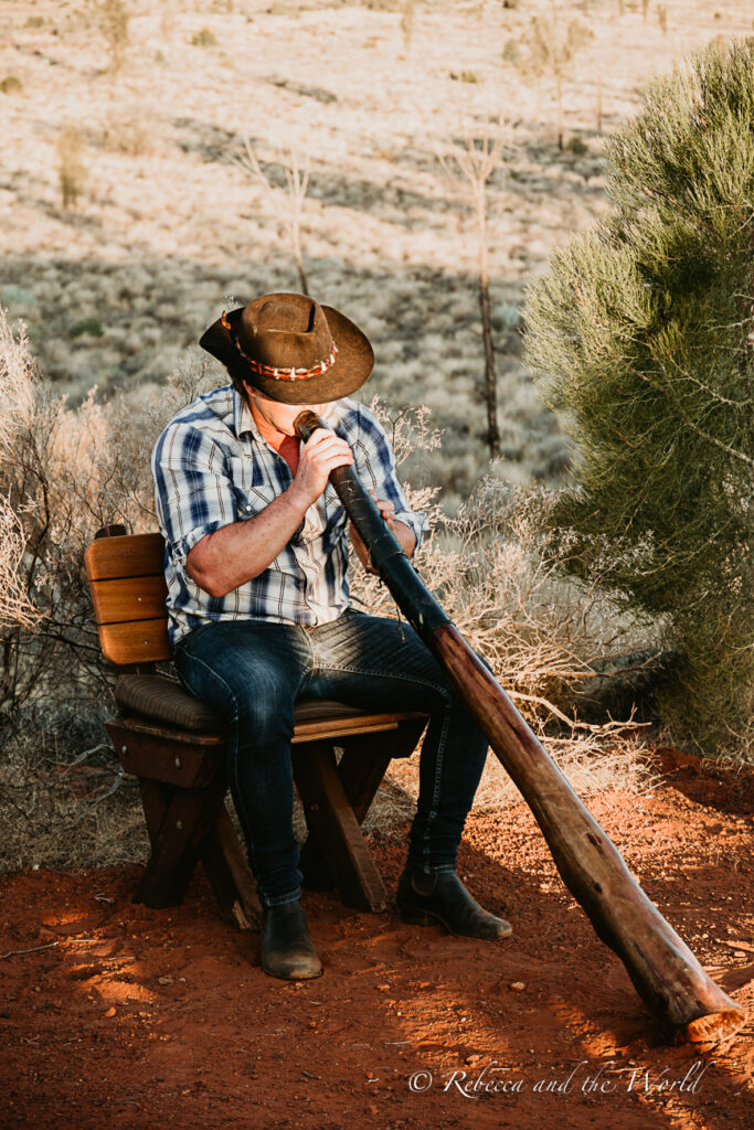 A person seated on a wooden chair outdoors, dressed in a blue checked shirt, jeans, and brown boots, wearing a brown hat. They are playing the didgeridoo, a traditional Australian wind instrument. The setting is a dusty, red earth landscape with sparse greenery in the background. One of the best things to do at Uluru is splash cash on one of the stunning dinners under the stars, such as Tali Wiru.