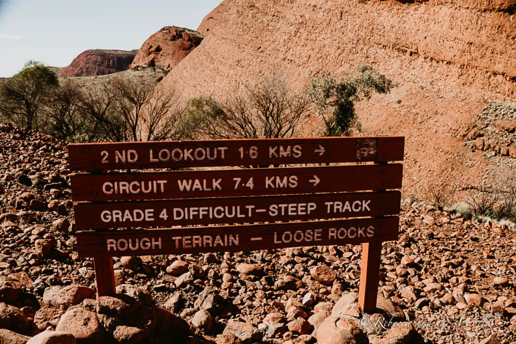 A wooden sign indicating directions and distances to various points of interest at Kata Tjuta, including a lookout and a circuit walk, with a backdrop of scattered rocks and sparse desert vegetation. The Valley of the Winds Trail is one of the best things to do near Uluru, in Kata Tjuta.