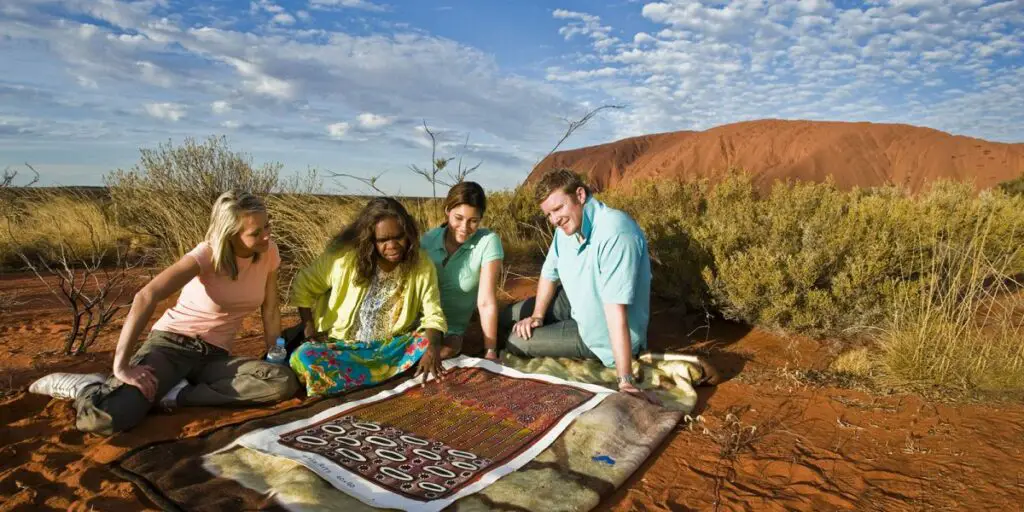 Four people sit on the ground at Uluru, engaging with a local Anangu guide who is sharing stories and cultural insights while presenting traditional indigenous artwork.