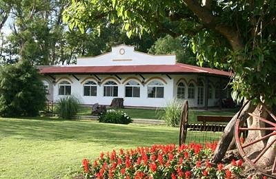 A white ranch-style house with a red-tiled roof, surrounded by greenery and a wooden wagon wheel, under a clear sky. The property is Estancia Don Manuel, a Buenos Aires estancia you can stay at.