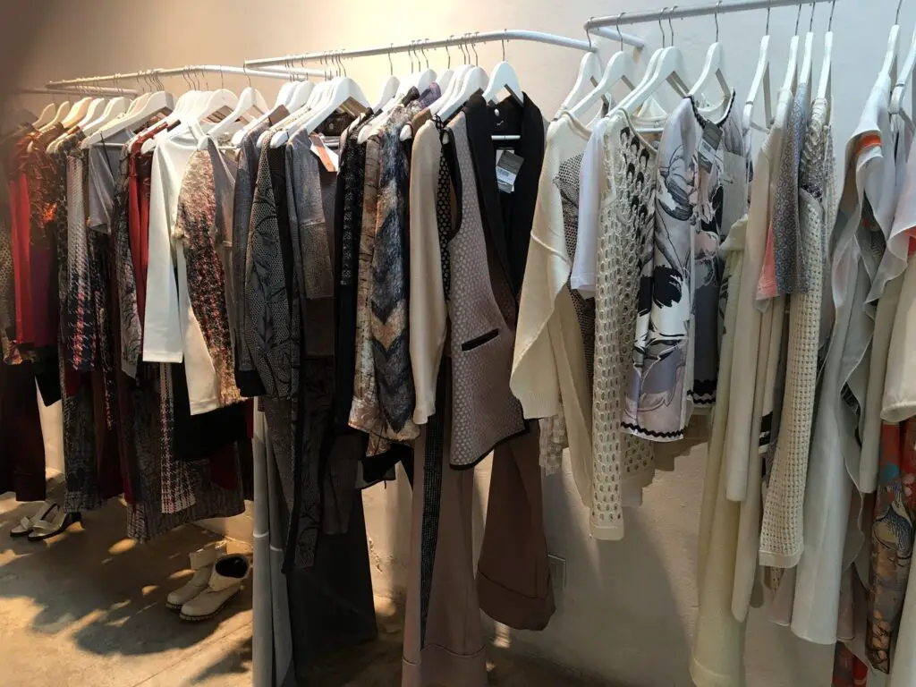 A variety of women's clothing hangs on racks in a retail store. The garments include coats, dresses, and knitwear, featuring an assortment of patterns and textures. Footwear is visible on the floor beneath the garments.