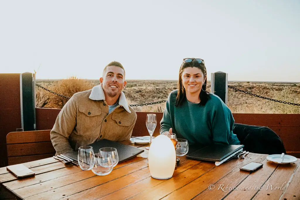 Two individuals - the author and her husband - seated at a wooden table outdoors, smiling and facing the camera, with wine glasses and a bottle on the table, and a flat, arid landscape in the background.