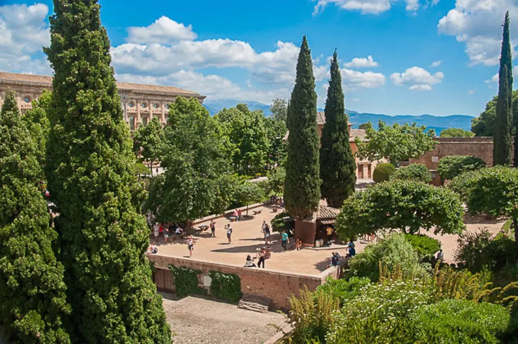 Aerial view of a historic courtyard surrounded by trees, with people walking and sitting in the shade, against a backdrop of a grand building and mountains. This is the stunning Alhambra in Granada, Spain.