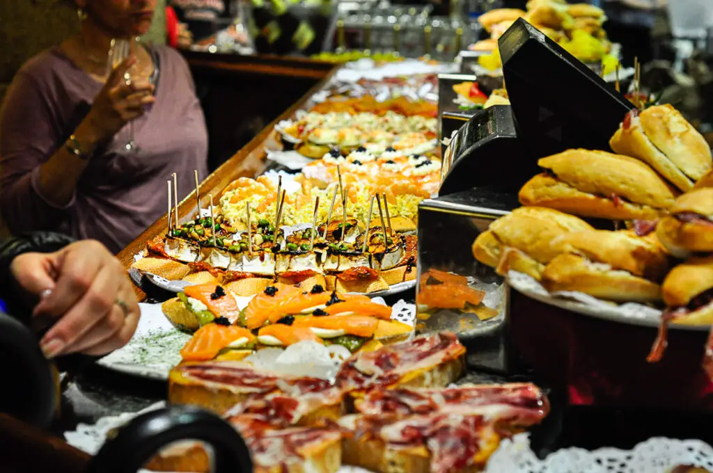 Busy indoor restaurant with an array of colourful tapas and sandwiches displayed, and patrons browsing and enjoying food. Pintxos in San Sebastian are part of the culture of the city and a must-try when you visit.