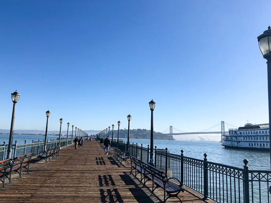 Wooden pier with street lamps and benches, overlooking a body of water with a bridge in the distance and a docked ferry to the right. This is San Francisco.