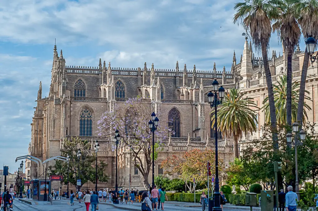 Gothic-style cathedral with flying buttresses and intricate facades, framed by tall palm trees under a blue sky with scattered clouds. The cathedral is in Seville, Spain.