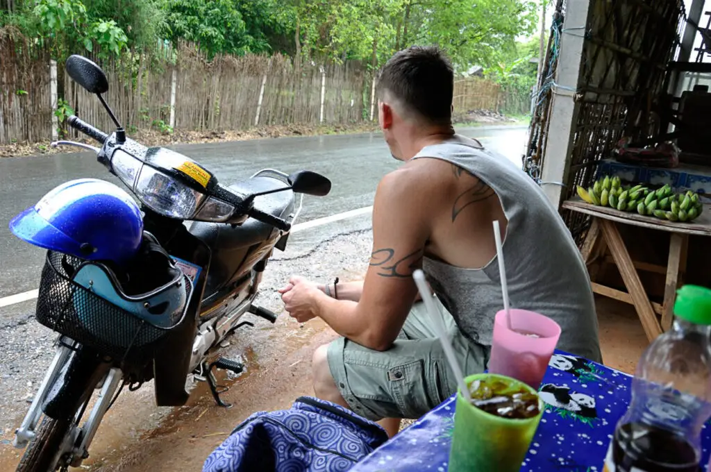 A man - the author's husband - sitting at an outdoor roadside stall with a motorbike parked in the foreground, surrounded by tropical vegetation. This was on a trip to Thailand when the rainy season was just about to start - it's important to know the main seasons when you plan a trip.