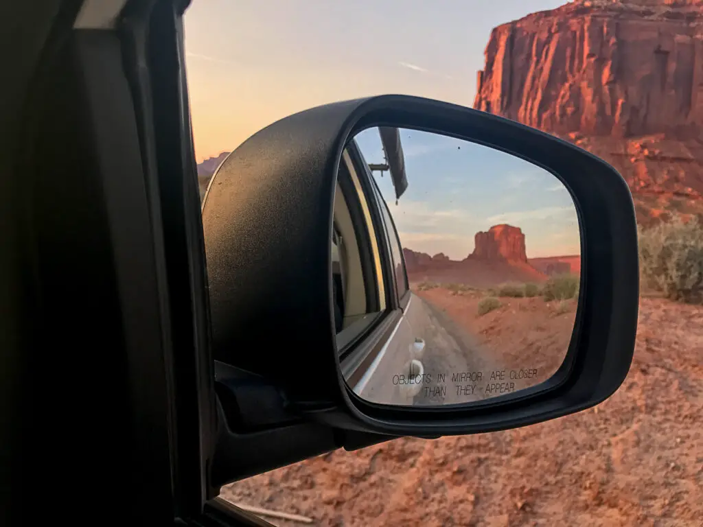 Car's side mirror reflecting a desert road with rock formations in the distance, text on the mirror reads 'Objects in mirror are closer than they appear.