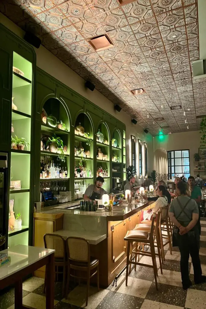 The warmly lit bar at Via Triozzi in Lower Greenville, Dallas, featuring elegant green cabinetry, a marble countertop, and an intricate tin ceiling. A bartender prepares drinks while guests enjoy the romantic ambiance of this intimate Italian restaurant.