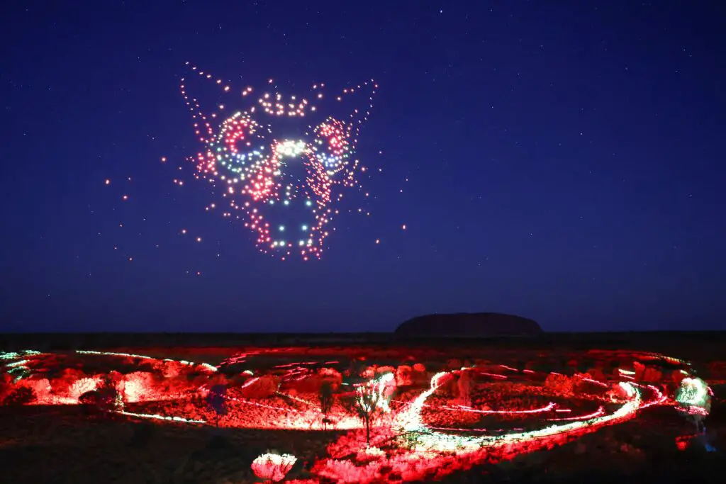 A night scene showcasing a light installation at Uluru, with patterns of bright red and white lights on the ground and a dazzling configuration of lights in the sky above. Wintjiri Wiru - Kurpany the devil dog. Photo by Getty Images for Voyages Indigenous Tourism Australia. Acknowledgement - Anangu share the Mala story, from Kaltukatjara to Uluru, through a drone, sound and light show designed and produced by RAMUS.