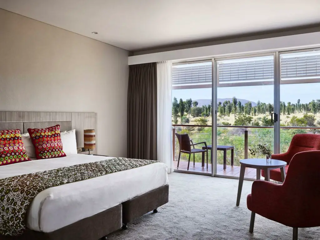 An interior view of a modern hotel room with a large window offering a panoramic view of a desert landscape. The room is furnished with a bed adorned with colorful pillows, a wooden headboard, and a seating area with a red armchair and a small table. Desert Gardens Hotel is a great Uluru accommodation option if you want views of Uluru - it's the only hotel with views of Uluru.