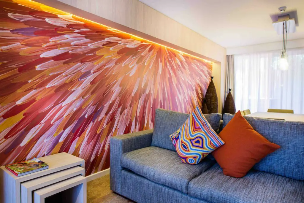 A colorful abstract painting adorning a wall in a hotel lounge area, which features a grey sofa with patterned cushions and a small wooden coffee table. Warm lighting enhances the artwork's vivid red, yellow, and orange hues. One of the Emu Walk Apartments at Uluru, a great accommodation option near Uluru.