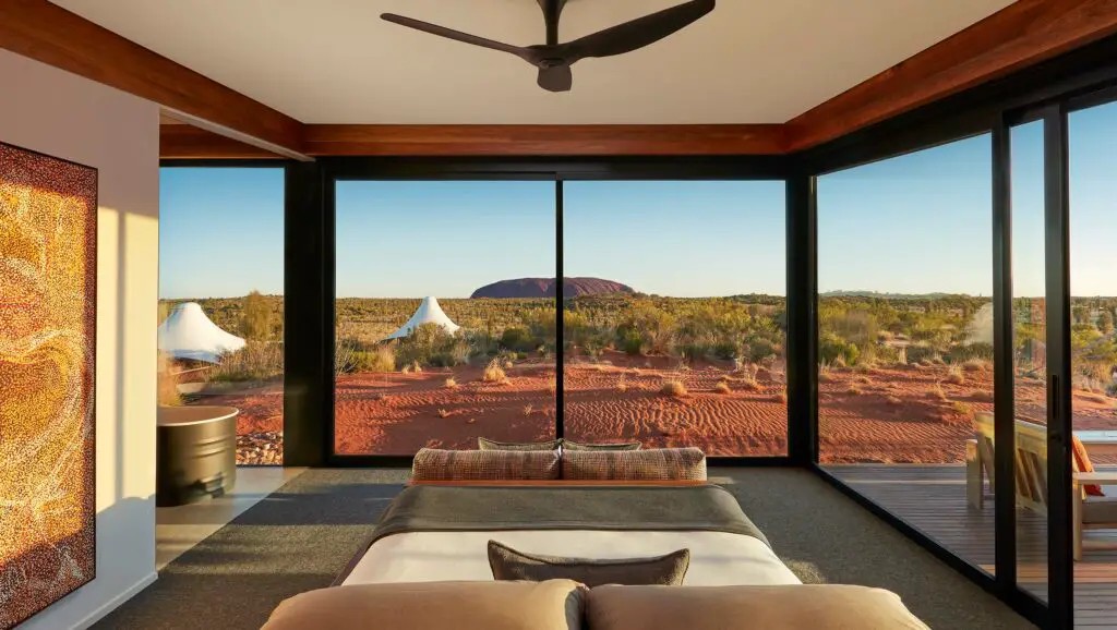 A luxurious room with large, floor-to-ceiling windows offering a stunning view of Uluru and the desert expanse. The room's decor includes a bed with a neutral bedspread, a ceiling fan, and wall art that complements the natural tones of the environment. Longitude 131 is the most expensive and exclusive Uluru accommodation, with luxury tents with views of Uluru.