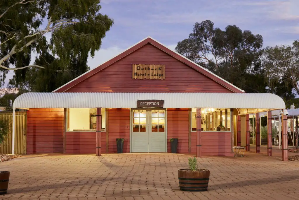 The entrance to a rustic-looking hotel with a red facade and 'Outback Hotel Lodge' signage above the door. The building has a corrugated iron roof, and the reception area is visible through the front glass doors. The Outback Hotel & Lodge is the most affordable place to stay if you're wondering where to stay near Uluru.