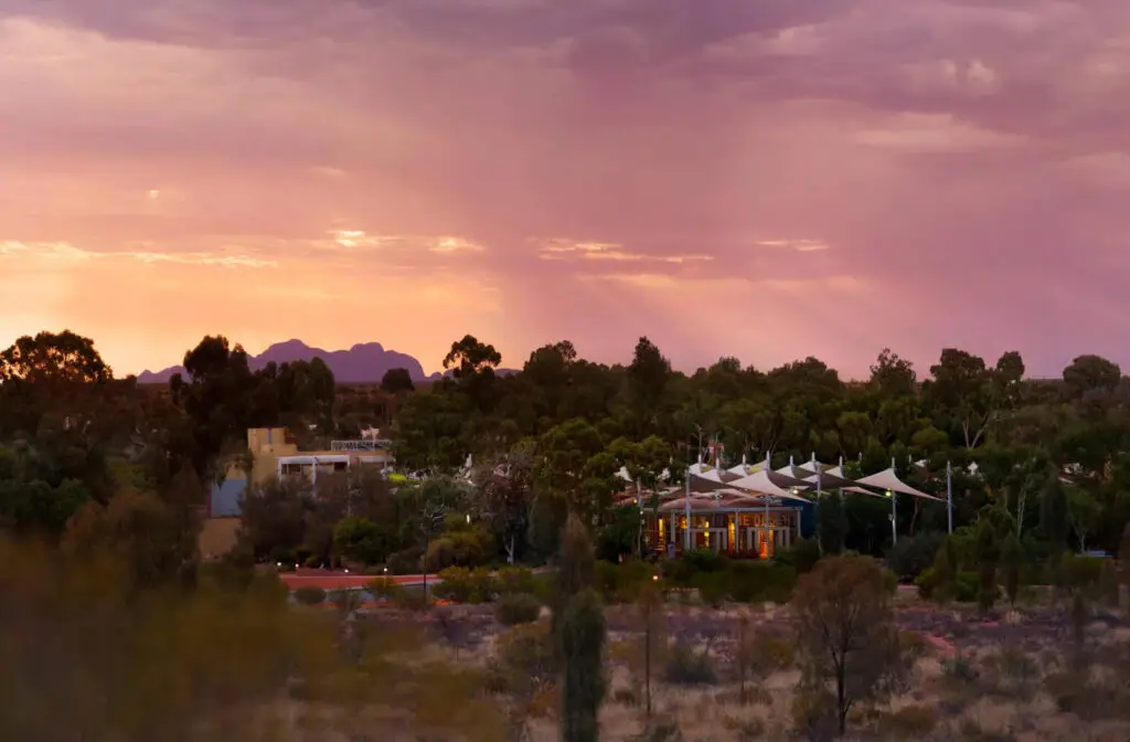 A twilight scene showcasing a resort with tents and low-rise buildings nestled among natural vegetation. The background features a soft, pink and purple sky with rays of sunlight filtering through the clouds, highlighting the distant silhouette of the Kata Tjuta rock formations. Sails in the Desert Hotel, a 5-star hotel near Uluru in Australia.